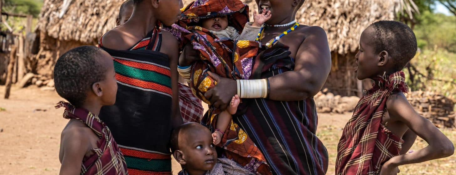 A woman holding a baby stands with several children outside thatched homes, Yaeda-Eyasi, Tanzania.