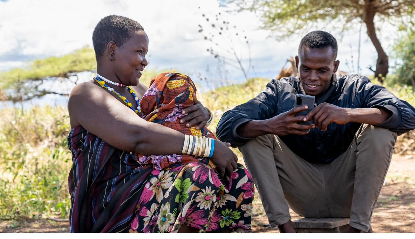 A woman holding a baby sits beside a fieldworker with a phone, in conversation, Yaeda-Eyasi, Tanzania.